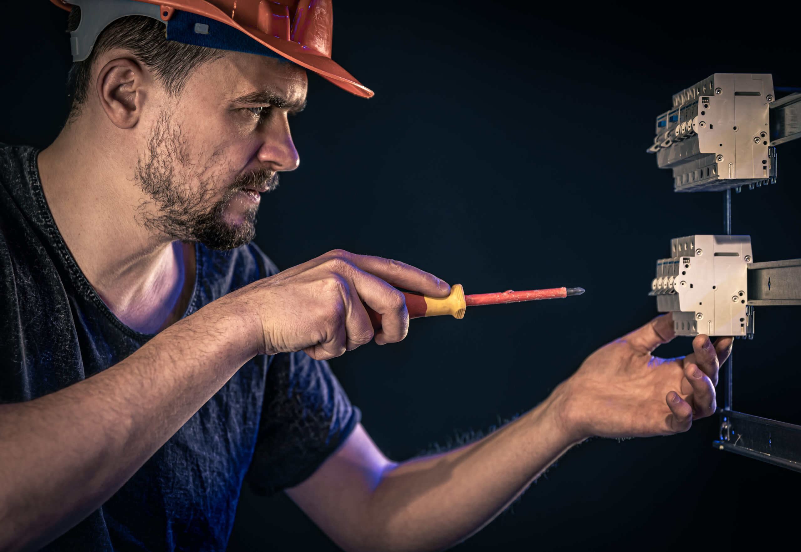A male electrician in a protective helmet works in a switchboard with an electrical connecting cable.