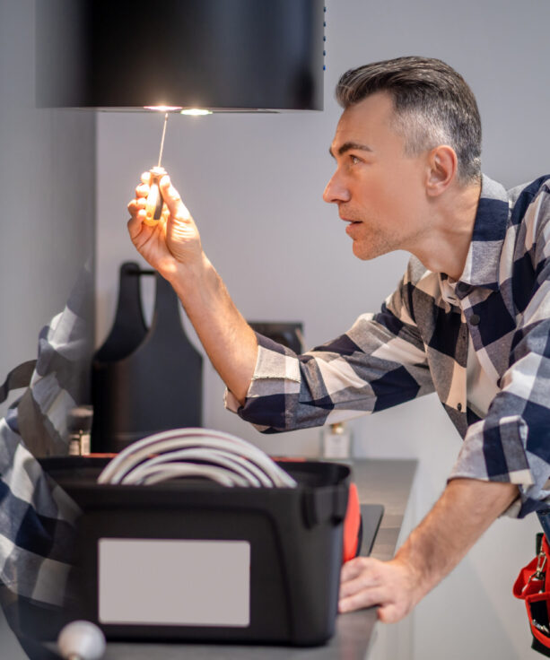 Renovation. Profile of concentrated middle aged man in plaid shirt touching screwdriver to lamp on kitchen range hood in modern kitchen at home